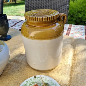 Könnte beinhalten: Eine weiße Keramik-Dose mit braunem Deckel und einem braunen Band um die Mitte. Die Dose hat einen Griff an der Seite. Die Dose steht auf einer braunen gewebten Oberfläche.
