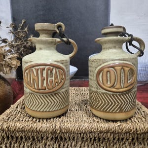 Peut inclure: Deux bouteilles à condiments en céramique, l'une avec l'inscription "VINEGAR" et l'autre "OIL". Les bouteilles sont beiges avec des lettres marron et un motif en chevrons. Chacune a une poignée et un bouchon attaché avec un cordon noir.