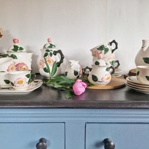 May include: A collection of vintage floral teacups, saucers, teapots, and a creamer. The white ceramic pieces are adorned with pink and yellow flower designs and green leaves. The set includes a serving tray, a sugar bowl, and a pitcher.