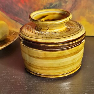 May include: A handcrafted ceramic jar with a lid. The jar features a cylindrical body and a rounded top with a small opening. The exterior is painted with horizontal bands of yellow and brown, creating a textured, earthy appearance. The jar is likely used for storage or decoration.