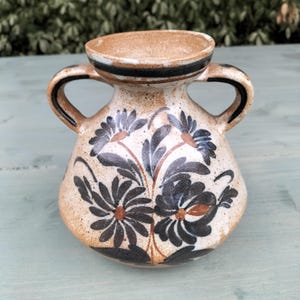 Peut inclure: Un petit vase en céramique brun avec deux anses et un motif floral. Le vase est décoré de fleurs et de feuilles noires et brunes.