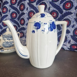 May include: White ceramic coffee pot with a blue floral design. The pot has a lid and a spout. The pot is sitting on a dark brown wooden surface.