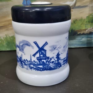 Puede incluir: Un tarro de cerámica blanco y azul con tapa azul oscuro. El tarro presenta una ilustración azul y blanca de un molino de viento, casas y un paisaje. El tarro tiene forma cilíndrica.
