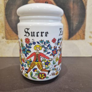 May include: White ceramic sugar jar with a lid. The jar has a colorful folk art design featuring three people dancing around a floral pattern. The jar is labeled "Sucre" in black lettering.