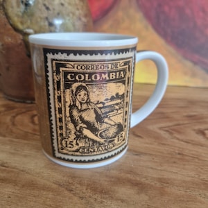 May include: A ceramic mug featuring a vintage postage stamp design from Colombia. The stamp illustration depicts a woman and the text "CORREOS DE COLOMBIA" in black. The mug has a white handle and rim, with a brown and tan background.