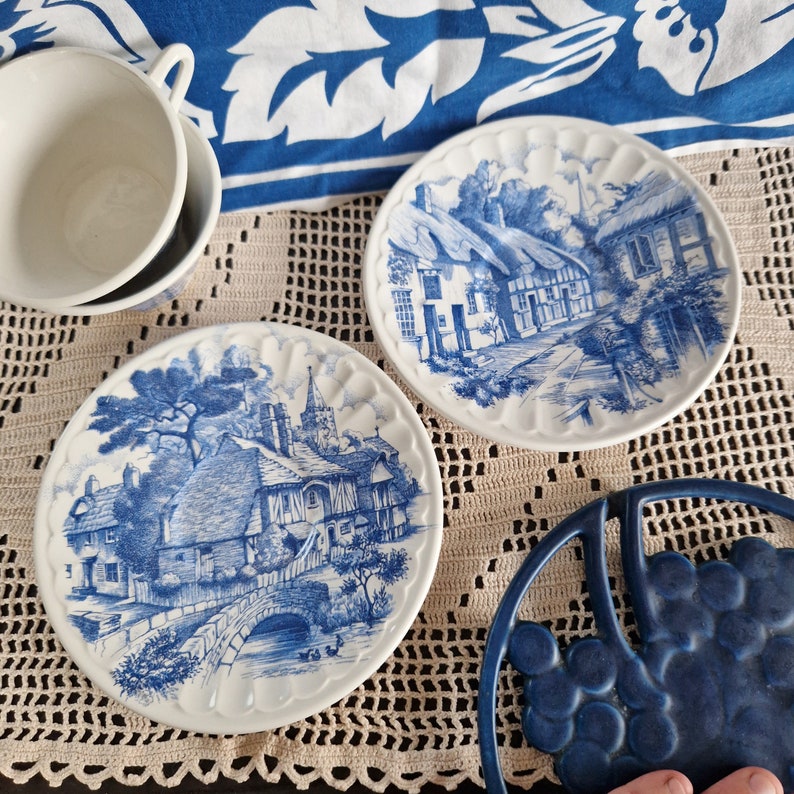 K&ouml;nnte beinhalten: Zwei blaue und wei&szlig;e Keramikteller mit einem Cottage-Motiv. Die Teller liegen auf einer wei&szlig;en H&auml;keldeckchen.