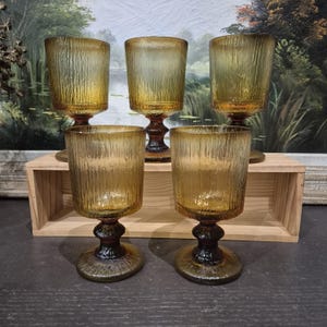 May include: Five amber-colored vintage goblets with textured glass. The goblets have a short stem and a wide base. The glass has a vertical ribbed pattern. The goblets are displayed on a wooden shelf against a backdrop of a landscape painting.