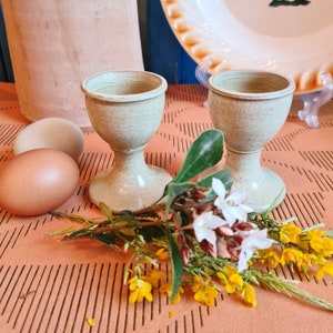 以下が含まれることがあります： 薄い緑色の斑点のある仕上げのセラミック製の卵カップが2つ。カップは茶色の表面に置かれ、その前に花が飾られています。