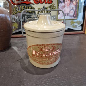 May include: A beige ceramic jar with a lid and a metal wire handle. The jar has a label that reads "Win Schuler Restaurant Bar-Scheeze Snappy Snack Spread - Imitation Cold Cheese Spread".