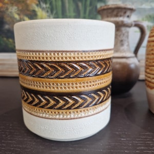 Puede incluir: Un macetero de cerámica blanca con patrones geométricos marrones y dorados. El macetero tiene una superficie texturizada y una forma redondeada.