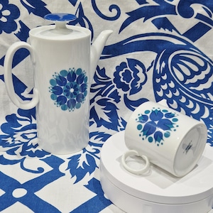 Puede incluir: Un juego de cafetera y taza de cerámica blanca con un diseño floral azul y verde. La cafetera tiene una tapa y un asa. La taza tiene un asa.