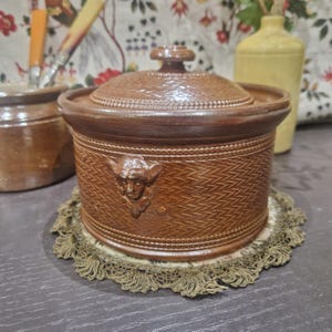 May include: A brown ceramic jar with a lid and a decorative design. The jar has a woven pattern and a small face on the front. The jar is sitting on a lace doily.