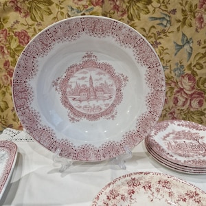 Peut inclure: Un ensemble d'assiettes en céramique vintage rose et blanche avec un motif floral et une image centrale d'un bâtiment. Les assiettes sont sur une nappe blanche.