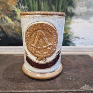 Peut inclure: Une tasse en céramique blanche avec un motif brun représentant un bouclier avec une épée et une hache croisées, entourées d'une couronne et d'une bannière avec le texte "SIGILLA DEI ET LIBERTAS".