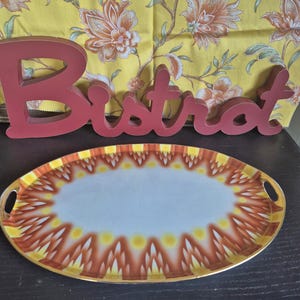 Op de afbeelding: Een ovaal dienblad met een wit midden en een decoratieve rand met herhalende oranje en gele vlamachtige patronen. Het dienblad heeft geïntegreerde handgrepen aan beide zijden en een goudkleurige rand. Achter het dienblad staat een rood houten bord met het woord Bistro.