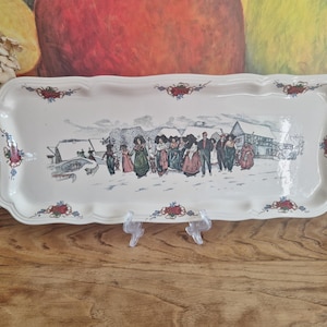 Puede incluir: Una bandeja rectangular de cerámica blanquecina con borde festoneado. La bandeja presenta una ilustración detallada de un grupo de personas con trajes tradicionales en una escena de pueblo nevado. Acentos florales rojos y azules decoran los bordes.