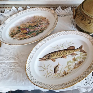 May include: Two oval ceramic serving platters with gold trim. One platter features a painted image of a pheasant in a forest setting. The other platter features a painted image of a fish in a pond setting.