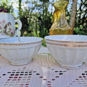 May include: Three white porcelain bowls with gold trim and a fluted design. The bowls are sitting on a white crocheted tablecloth.