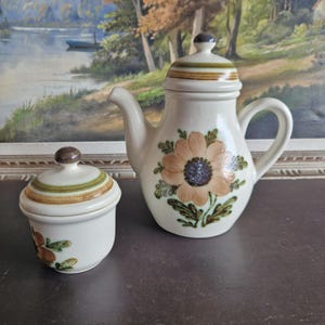 May include: A vintage ceramic teapot and sugar bowl set with a floral design. The teapot has a brown, green, and white floral pattern with a brown lid. The sugar bowl has a matching floral pattern with a brown lid.