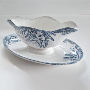 Op de afbeelding: Een witte porseleinen sauskom met een blauw bloemenmotief. De kom heeft een gegolfde rand en staat op een bijpassend schoteltje.