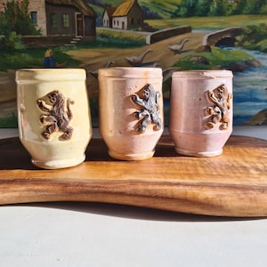 Peut inclure: Trois mugs en céramique avec un motif de lion. Les mugs sont dans des tons de rose et de jaune. Les mugs sont posés sur une surface en bois.