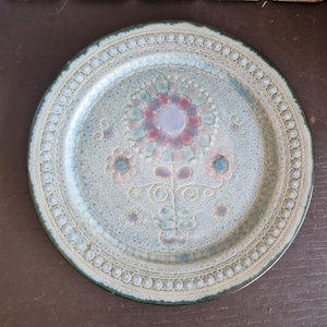 May include: A round ceramic plate with a floral design. The plate features a light blue background with a central flower in shades of purple and pink. The rim has a dotted pattern and a green edge.