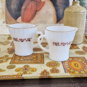 Peut inclure: Deux crémiers blancs avec des bords dorés et des motifs floraux. Les crémiers ont des anses et sont posés sur un plateau décoratif avec un motif orange et jaune. Un vase beige est à l'arrière-plan.