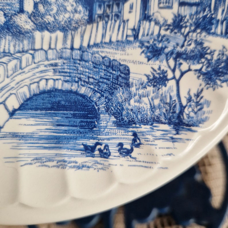 K&ouml;nnte beinhalten: Ein wei&szlig;er Keramikteller mit einem blauen und wei&szlig;en Design einer Br&uuml;cke &uuml;ber einen Fluss mit drei schwimmenden Enten. Der Teller hat einen gewellten Rand und ist perfekt zum Servieren von Snacks oder Vorspeisen.