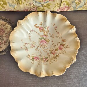 Peut inclure: Un bol en céramique de couleur crème avec un bord festonné et ondulé ainsi qu'un contour dégradé beige. Le centre présente un motif floral délicat avec des roses roses, de petites fleurs blanches, des feuilles vertes et un motif doré.