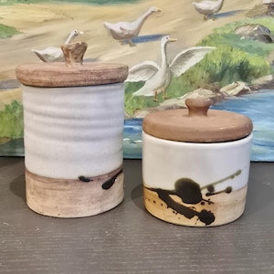 Peut inclure: Deux pots en céramique avec couvercles. L'un est grand et cylindrique avec une base et un couvercle marron clair, et un corps blanc avec un motif texturé. L'autre est plus court, avec un corps blanc, une base et un couvercle marron et un motif éclaboussé noir.