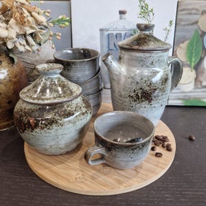 Op de afbeelding: Een keramisch theeservies met een gespikkeld grijs en bruin glazuur. De set bevat een theepot, een suikerpot met deksel, een kopje en een schotel. Het theeservies staat op een houten dienblad.