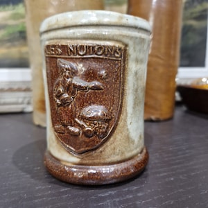 Peut inclure: Une tasse en céramique brune et blanche avec un motif brun d'une personne tenant un oiseau. La tasse porte le texte "LES NUTONS" sur le devant.