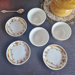 Peut inclure: Ensemble de vaisselle ancienne en porcelaine blanche avec une bordure décorative dorée, rouge et jaune. L'ensemble comprend de petites assiettes et des bols, ainsi qu'une cuillère en argent. Les plats sont disposés sur une surface sombre.