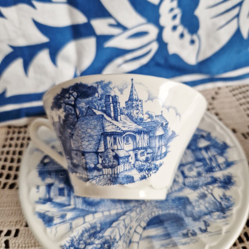 K&ouml;nnte beinhalten: Eine wei&szlig;e Teetasse und Untertasse mit einem blauen und wei&szlig;en Blumenmuster. Das Muster zeigt ein H&auml;uschen mit einer Kirche im Hintergrund.