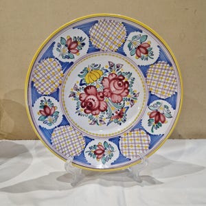 Puede incluir: Un plato de cerámica blanco con un borde azul y amarillo. El centro del plato presenta un diseño floral con flores rosas, rojas, amarillas y verdes. El borde está decorado con un patrón a cuadros.