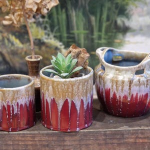 Peut inclure: Trois pots en céramique avec un glaçage rouge, blanc et brun. Les pots sont sur une surface en bois. Un pot contient une plante succulente.