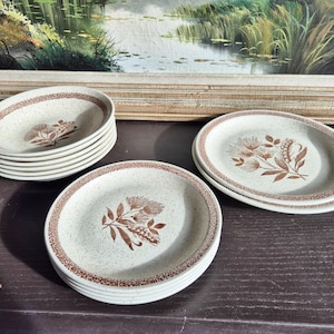 Op de afbeelding: Een set vintage steengoed servies met een gespikkelde beige afwerking en een bruin bloemmotief. De set bevat ovale borden en kommen, perfect om maaltijden te serveren. De borden hebben een bruine rand.