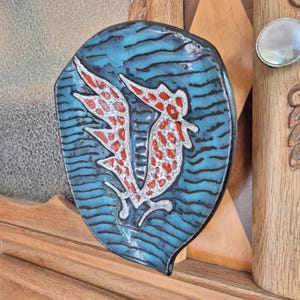 以下が含まれることがあります： シーホースのデザインが施された装飾用セラミックプラーク。プラークは、黒い波のような線が入った青色の背景と、赤いディテールが施された白いシーホースが特徴です。木製の表面に取り付けられています。