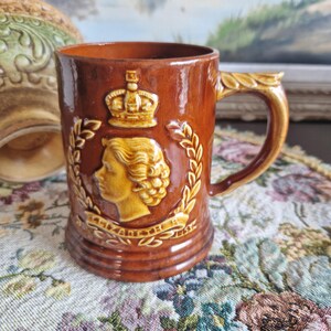 Peut inclure: Une tasse en céramique brune avec une image en relief de la reine Élisabeth II, une couronne et une couronne de laurier. La tasse a une anse et est décorée d'un glaçage doré.