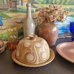 Peut inclure: Un beurrier en céramique avec un couvercle orné d'un motif marron et crème. Le plat est rond avec un petit bouton sur le dessus. D'autres articles incluent un vase, une bouteille, un verre et une assiette.