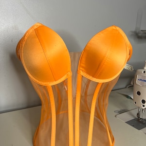 May include: A bright orange mesh corset with satin cups. The corset has vertical seams and a sweetheart neckline.