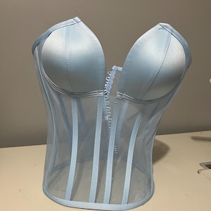 May include: A light blue corset with a sweetheart neckline. The corset features a sheer mesh overlay with vertical boning and a hook-and-eye closure down the front. The cups are made of a satin-like material.