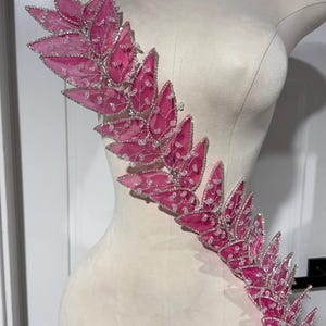 Könnte beinhalten: Dekorative, rosa-farbene, blattförmige Verzierung mit silberfarbenem Besatz und transparenten Perlenakzenten. Die Verzierung wird auf einer Schaufensterpuppe präsentiert, um ihr Design und ihre potenzielle Verwendung in Mode oder Handwerk hervorzuheben.