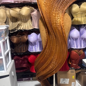 May include: A copper-colored, pleated fabric sculpture on a metal stand. The sculpture is draped over a mannequin, with a variety of corsets in the background. The corsets are in shades of pink, beige, brown, and purple.