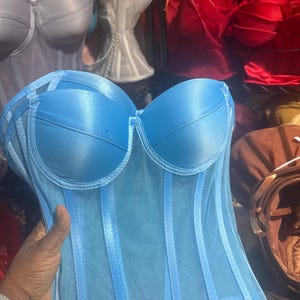 May include: A light blue corset with a satin finish and sheer overlay. Vertical boning and a structured bust create a defined silhouette. Other corsets in pink, white, and red are visible in the background, suggesting a lingerie collection.