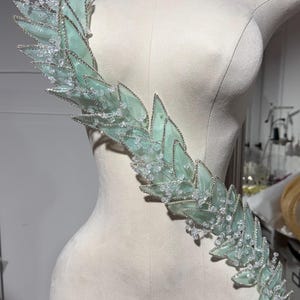 May include: A decorative shoulder piece with a leaf design. The leaves are a soft, pale green color, outlined with silver beading and adorned with clear, sparkling crystals. The piece is displayed on a mannequin.