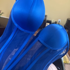 May include: A close-up of two royal blue corsets. The corsets have a structured, padded bust and sheer mesh panels with vertical support straps. The fabric has a satin sheen. The corsets are in a home setting.
