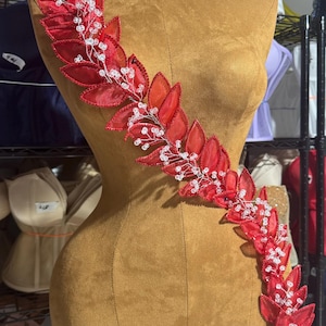 Puede incluir: Una aplicación floral roja y blanca con cuentas y un diseño de hojas. La aplicación está en un maniquí de tela marrón.