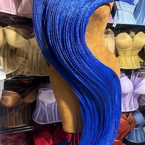 May include: A royal blue, pleated fabric draped over a mannequin. The fabric cascades down in vertical folds, creating a flowing, textured effect. The background features a variety of corsets in different colors, suggesting a lingerie or costume design theme.