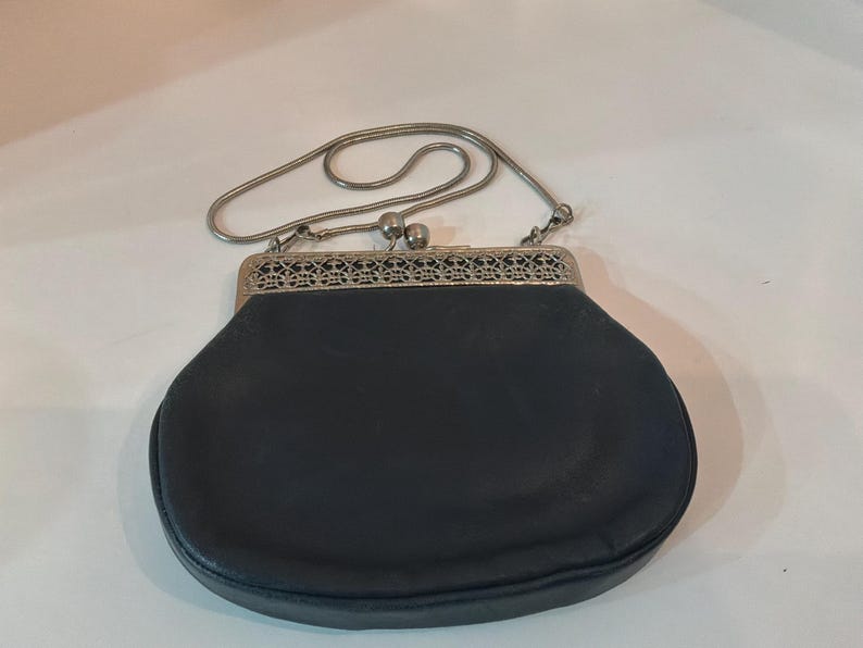 Pu&ograve; includere: Una borsa nera vintage con una tracolla a catena in metallo argentato. La borsa ha una forma arrotondata con una chiusura decorativa in metallo. La chiusura presenta un design traforato e intricato. La borsa &egrave; realizzata in un materiale scuro e liscio.